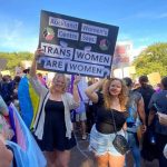 Kaitlin and Billie holding a sign at a rally that says "Auckland Women's Centre Says: TRANS WOMEN ARE WOMEN"