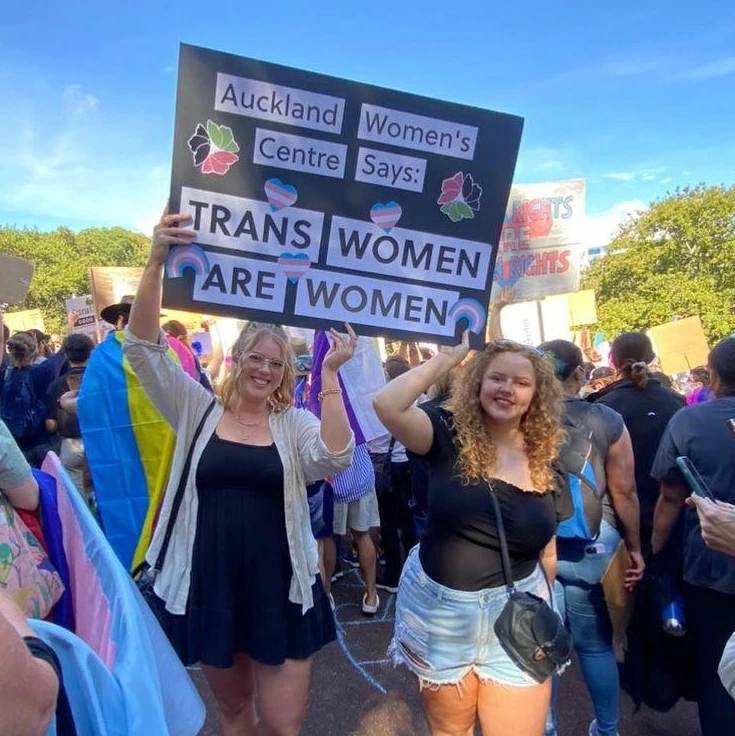 Kaitlin and Billie holding a sign at a rally that says "Auckland Women's Centre Says: TRANS WOMEN ARE WOMEN"