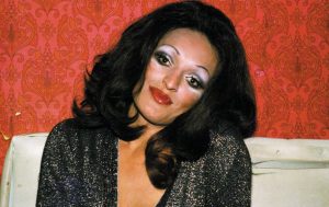 A trans woman in 1970s wearing bright make up and glamourous deep-v garment. 