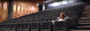 Tere Insley sitting in an auditorium with dark grey seats and light brown diamond-patterened walls