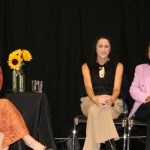 Stacey Morrison in warm brown dress with Dr Elana Curtis with a carved cream pendant and Lady Tureiti Moxon in a pink jacket, with sunflowers against a black background