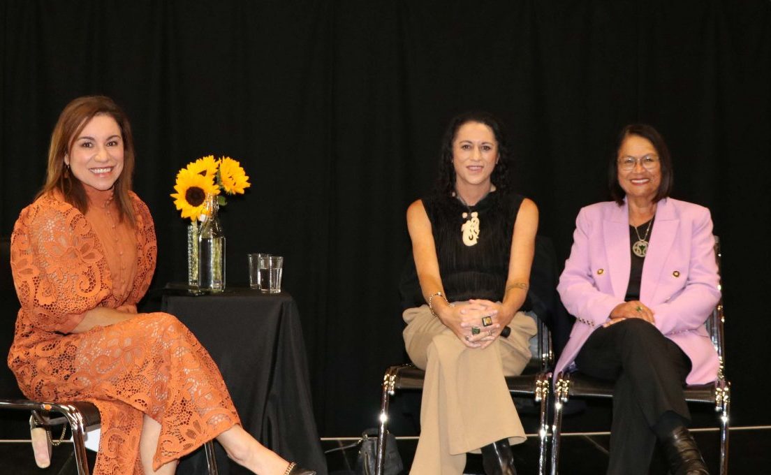 Stacey Morrison in warm brown dress with Dr Elana Curtis with a carved cream pendant and Lady Tureiti Moxon in a pink jacket, with sunflowers against a black background
