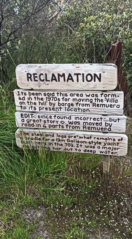 A sign made of multiple wooden planks, nestled in bush, saying “RECLAMATION It’s been said this area was formed in the 1970s for moving the villa on the hill by barge from Remuera to its present location. EDIT: since found incorrect… but a great story 😊 Was moved by road in 4 parts from Remuera.” Then more text about something else.
